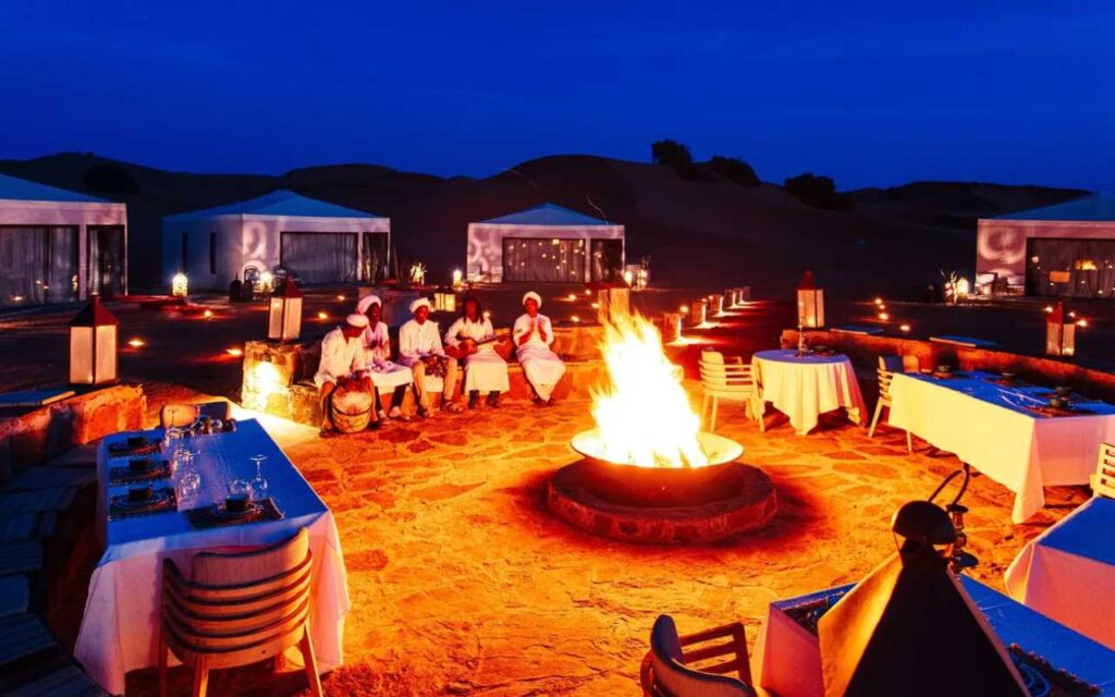 A firepit at night, surrounded by mobile safari tents, men sit round the fire playing instruments.